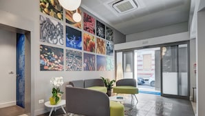 Un vestíbulo moderno con piso de baldosas, zona de estar con sillas verdes y grises, una planta en maceta y una pared decorada con diversas obras de arte enmarcadas.