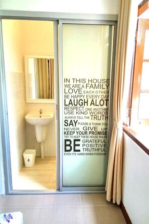 A bathroom with a sink, a mirror, and a glass door with a family house rules poster. - Banrublom (Rayong)