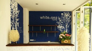 Uma área de recepção com parede azul, parede de tijolos brancos e balcão de madeira. Há uma placa com a inscrição "white at sea rayong" e uma citação de Voltaire.