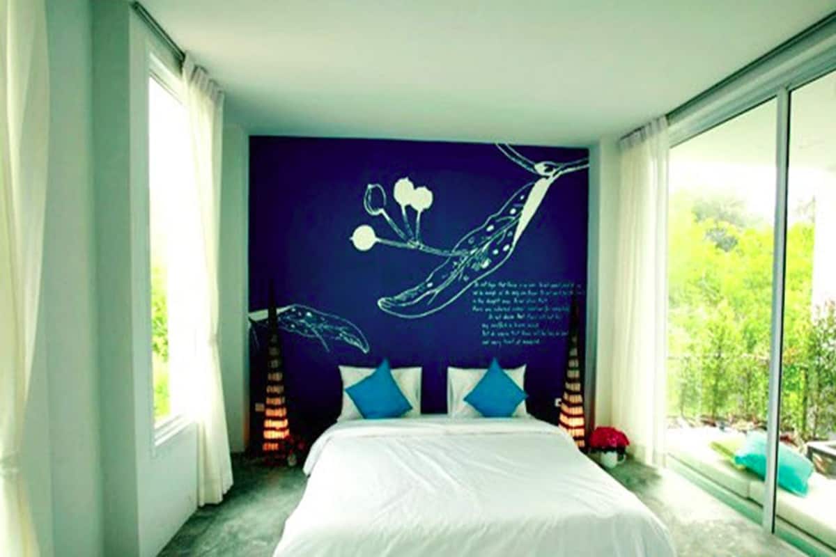 A bedroom with a large bed, a dark wall with a tree branch and text, and a view of greenery through a window.