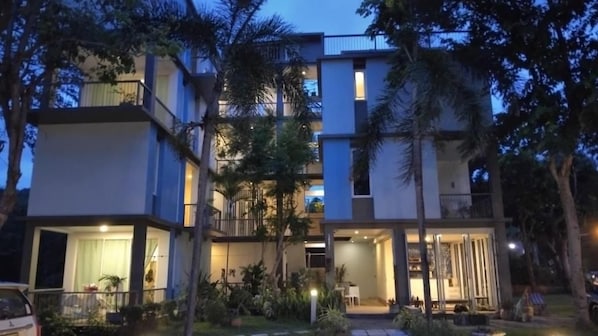 A multi-story residential building with balconies, a well-lit entrance, and a garden area.
