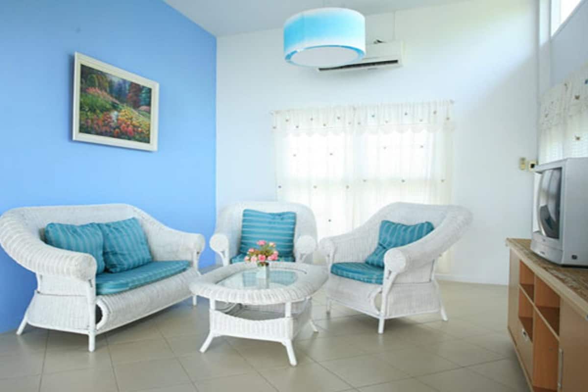 A living room with two white wicker sofas, a glass coffee table, a painting on the wall, a window with curtains, and a television on a wooden cabinet.