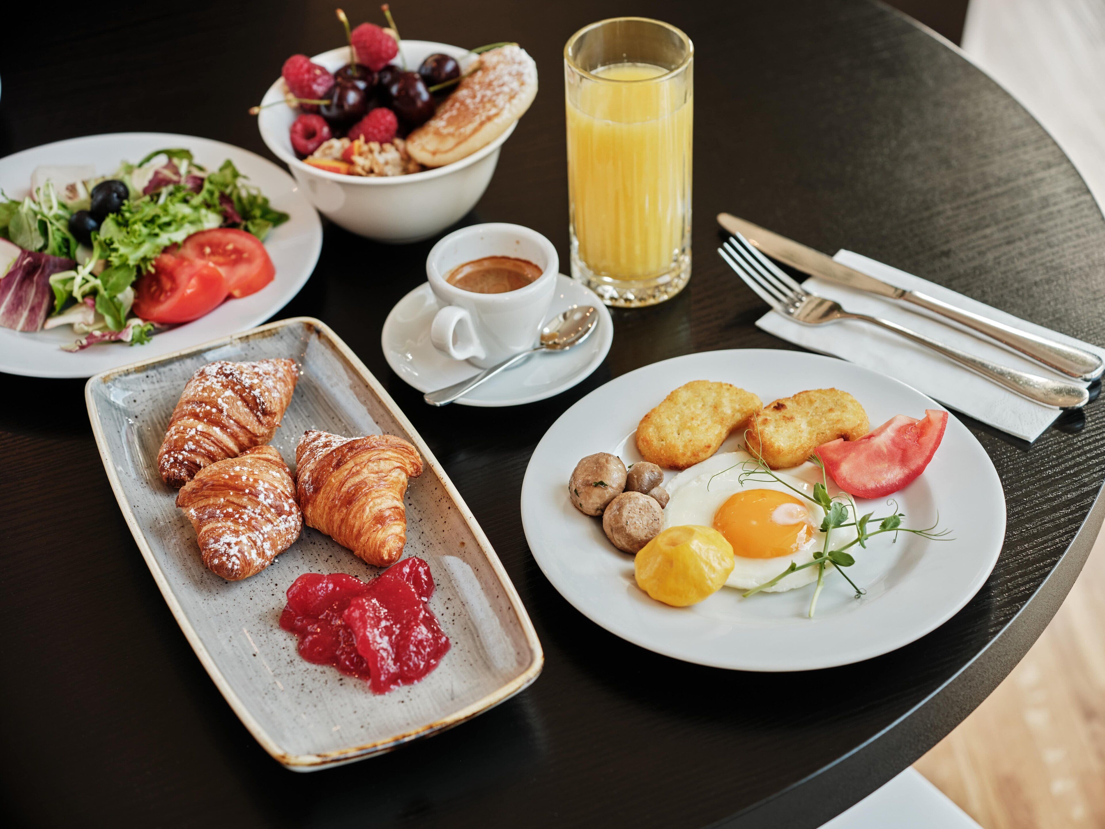 Aamiaispöytä, jossa on croissantteja, salaattia, hedelmiä, munakokkelia ja lasi appelsiinimehua.
