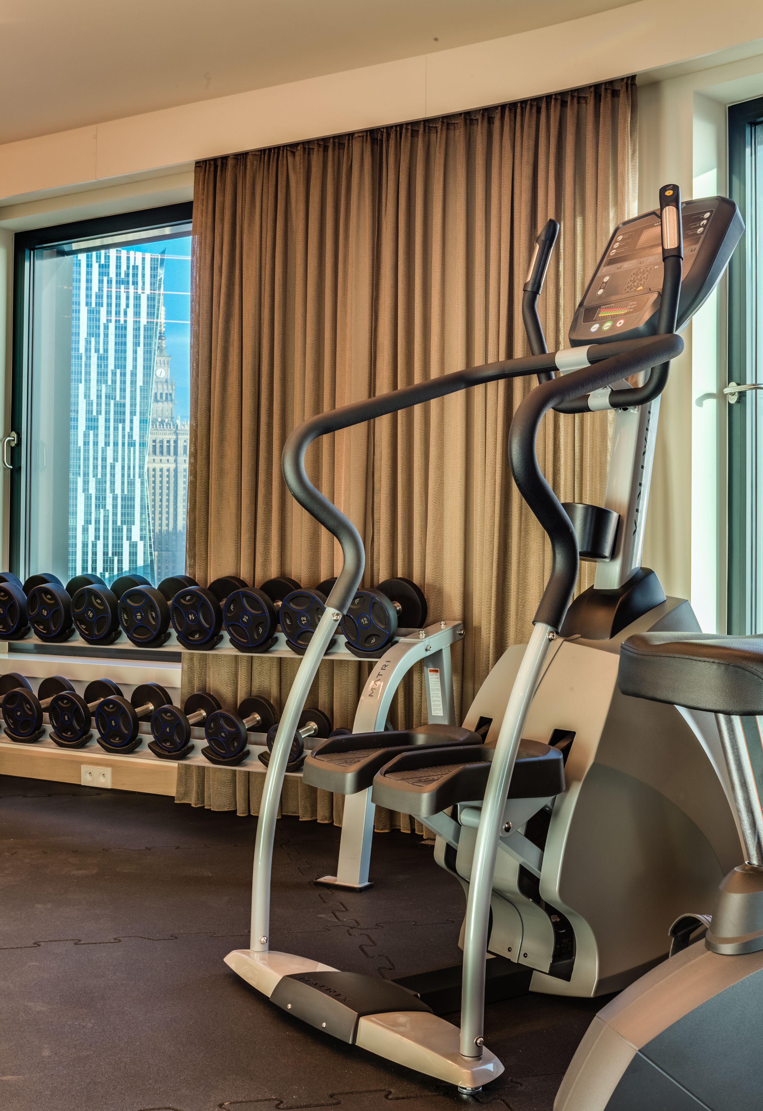 A gym with a treadmill, dumbbells, and a window with a cityscape view.