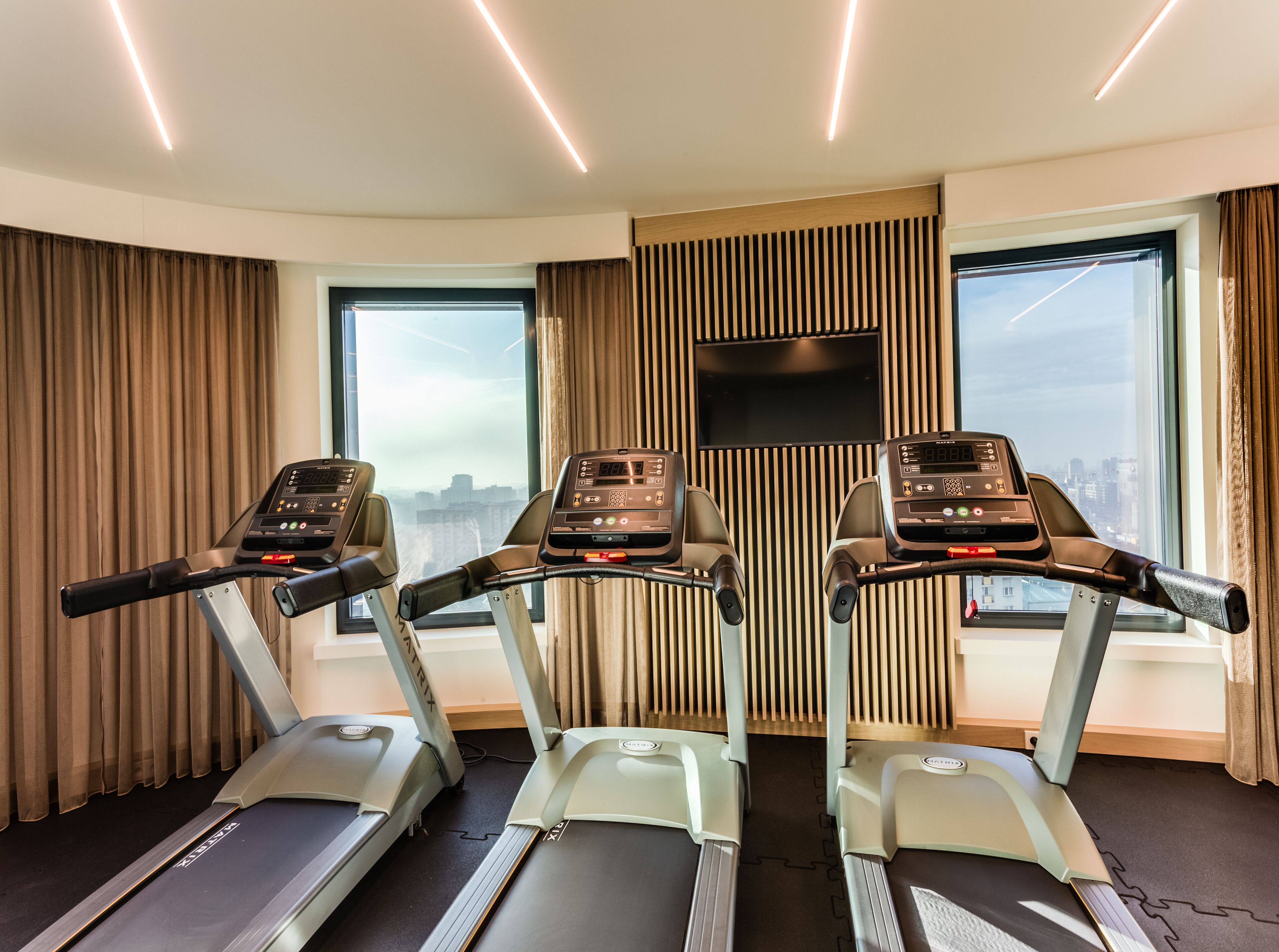 A hotel room with three treadmills aligned against a wall with a city view, a TV mounted above, and a patterned curtain.
