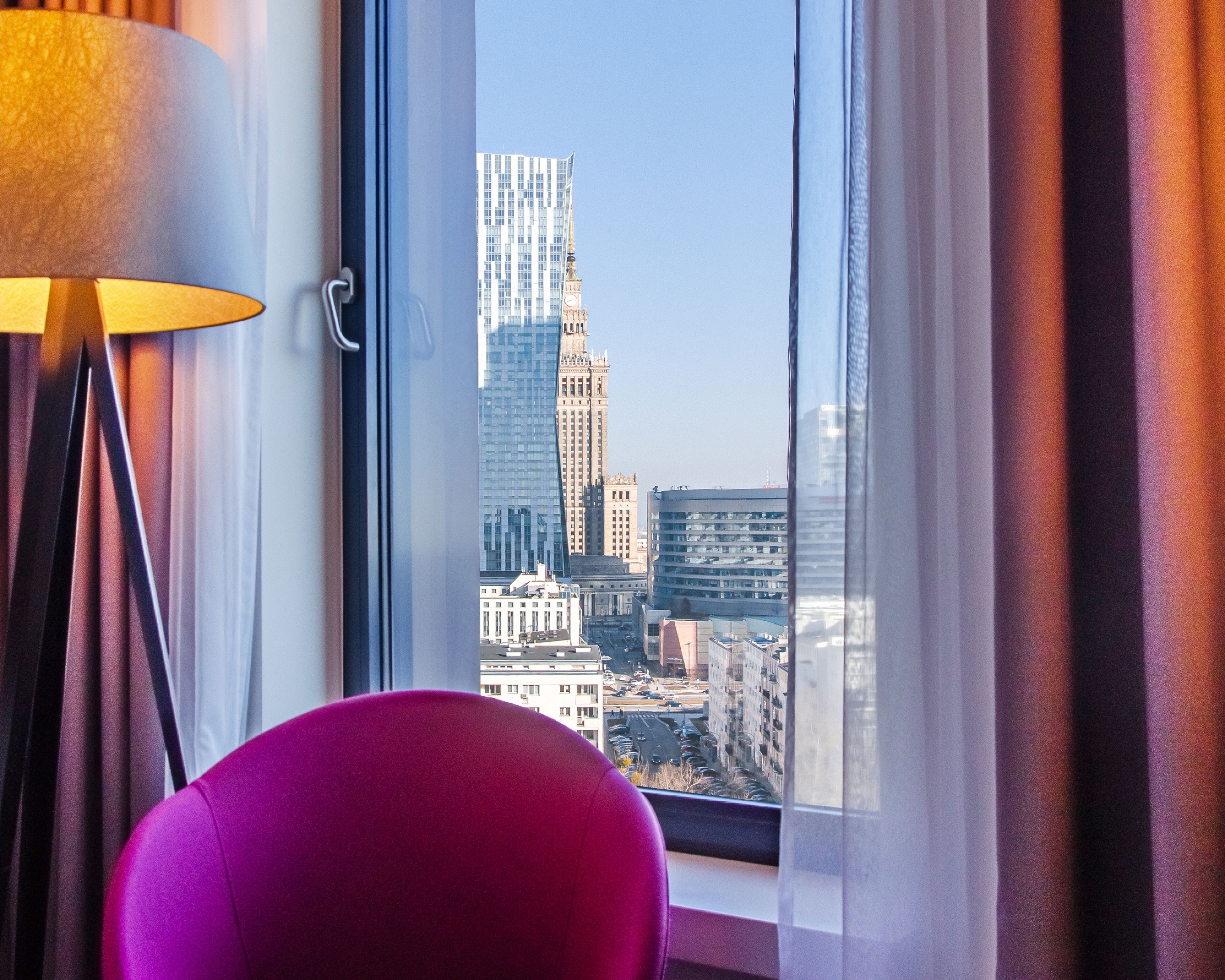 A room with a view of a cityscape through a window, a lamp, and a chair.