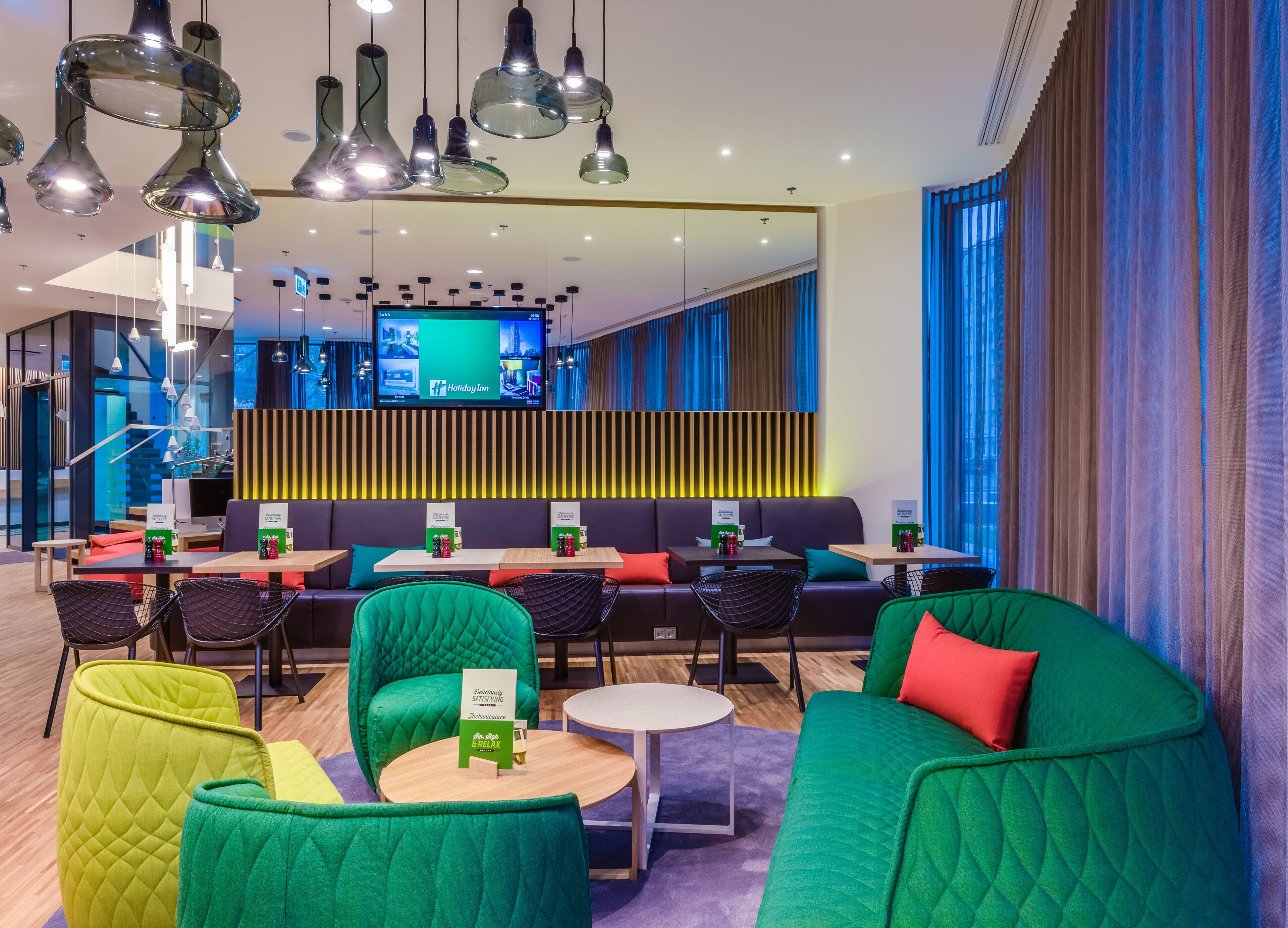 A modern hotel lobby with green and yellow seating, a TV screen displaying the Holiday Inn logo, and a wooden panel wall.
