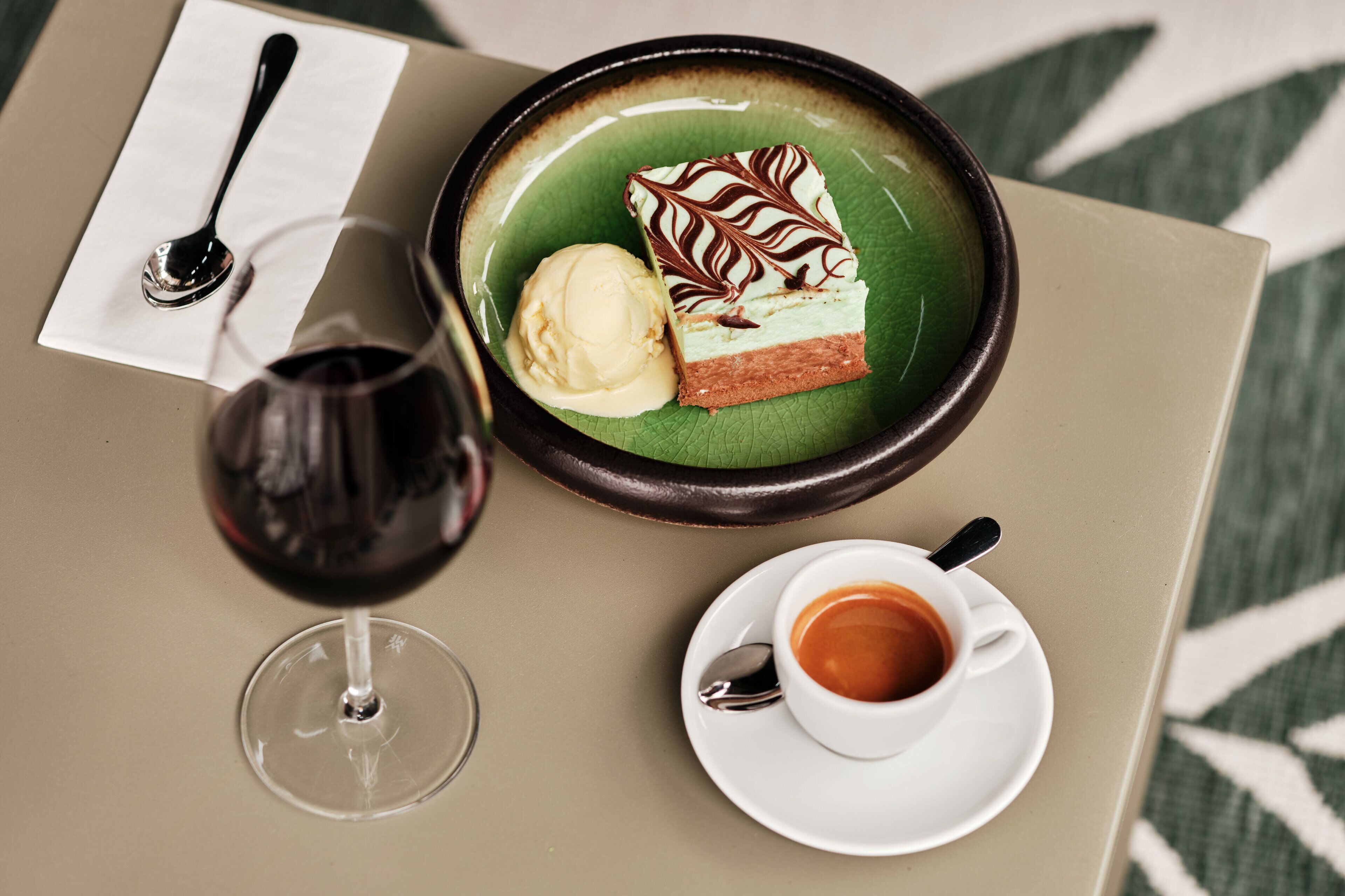 A dessert plate with a slice of cake and a scoop of ice cream, a glass of red wine, and a cup of coffee on a table.
