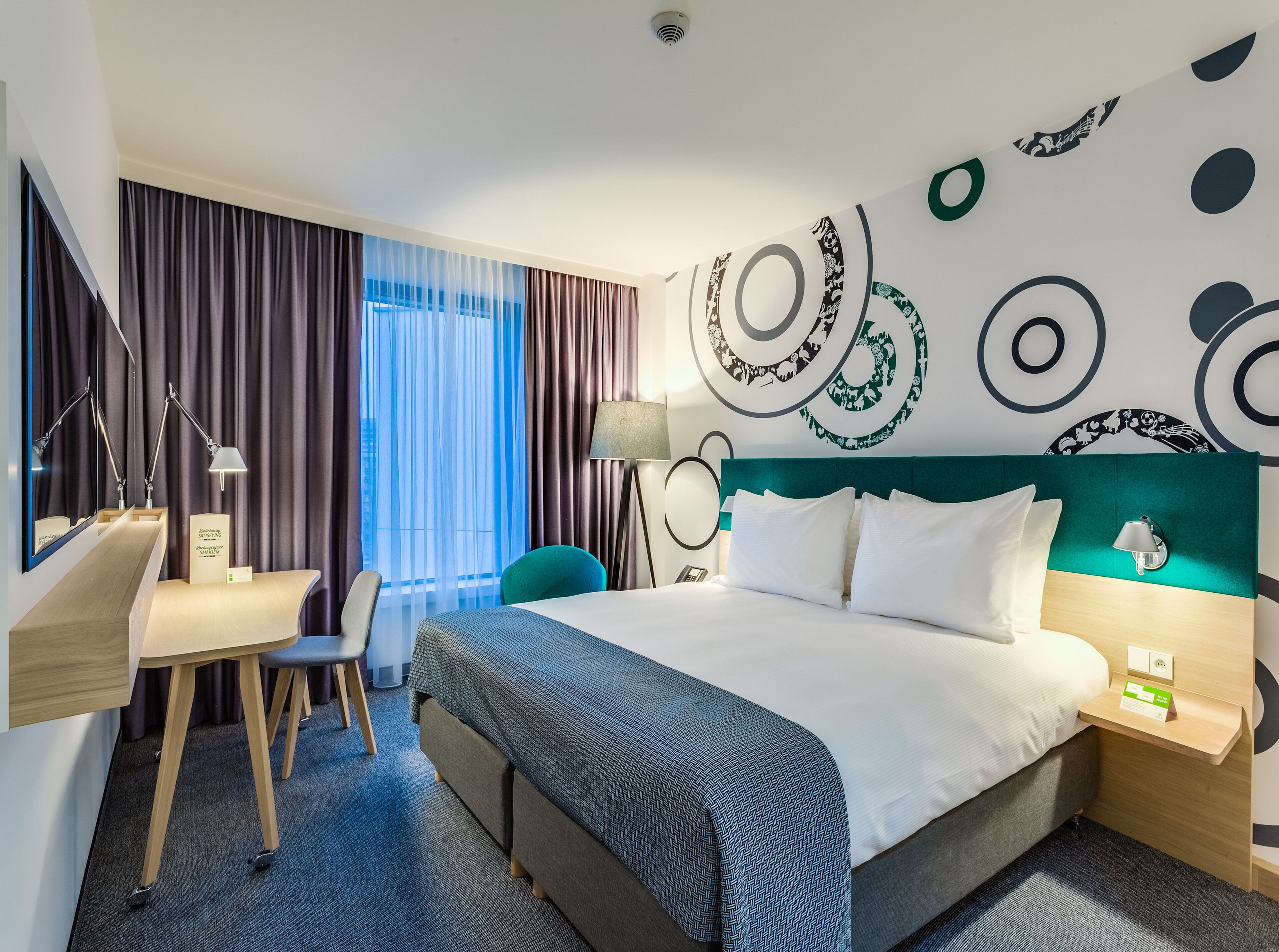 Habitación de hotel moderna con una cama grande, un escritorio, una silla y vistas de la ciudad a través de cortinas traslúcidas.