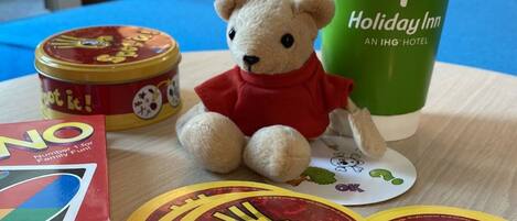 Una mesa con un osito de peluche, una caja etiquetada como “¡Número 1 para la Diversión Familiar!”, una lata con pegatinas de “Spot it!”, y una taza verde con el logotipo de “Holiday Inn”.