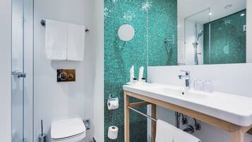 Een moderne badkamer met een glazen douchecabine, een wit toilet, een wastafel met spiegel en een wand betegeld met groene mozaïek.