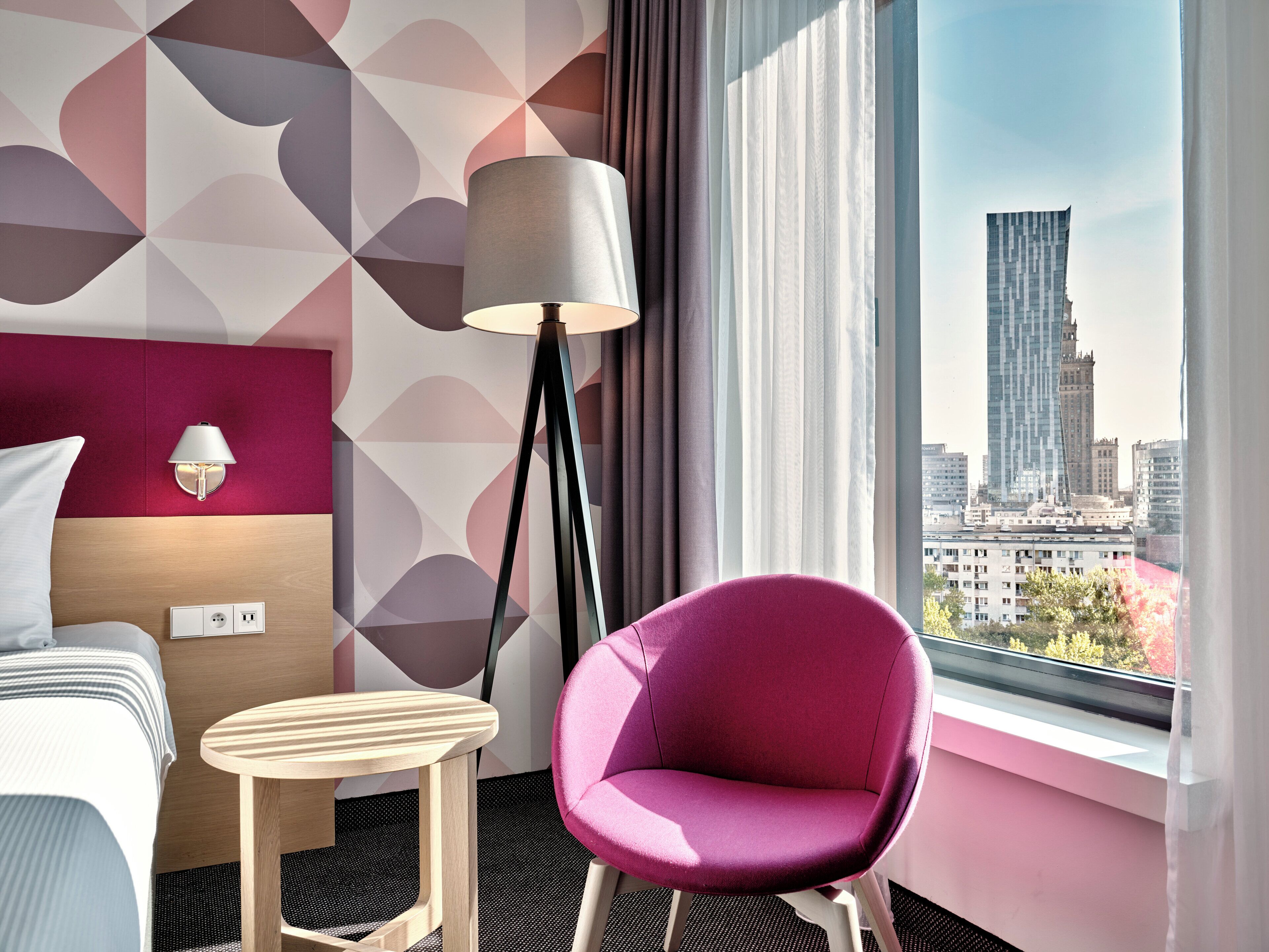 A hotel room with a modern design, featuring a bed, a small table, a chair, a lamp, and a large window offering a city view.