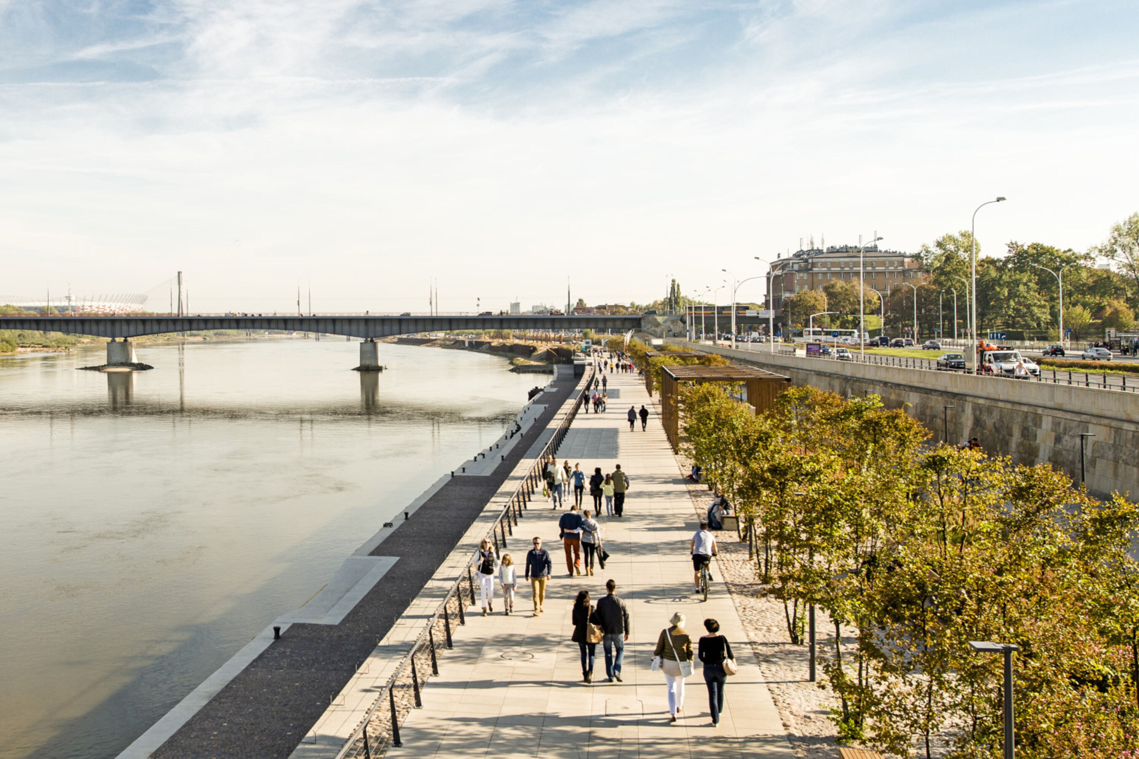 Een voetpad langs de rivier met mensen die lopen, een brug op de achtergrond en een stadsgezicht met gebouwen en bomen.