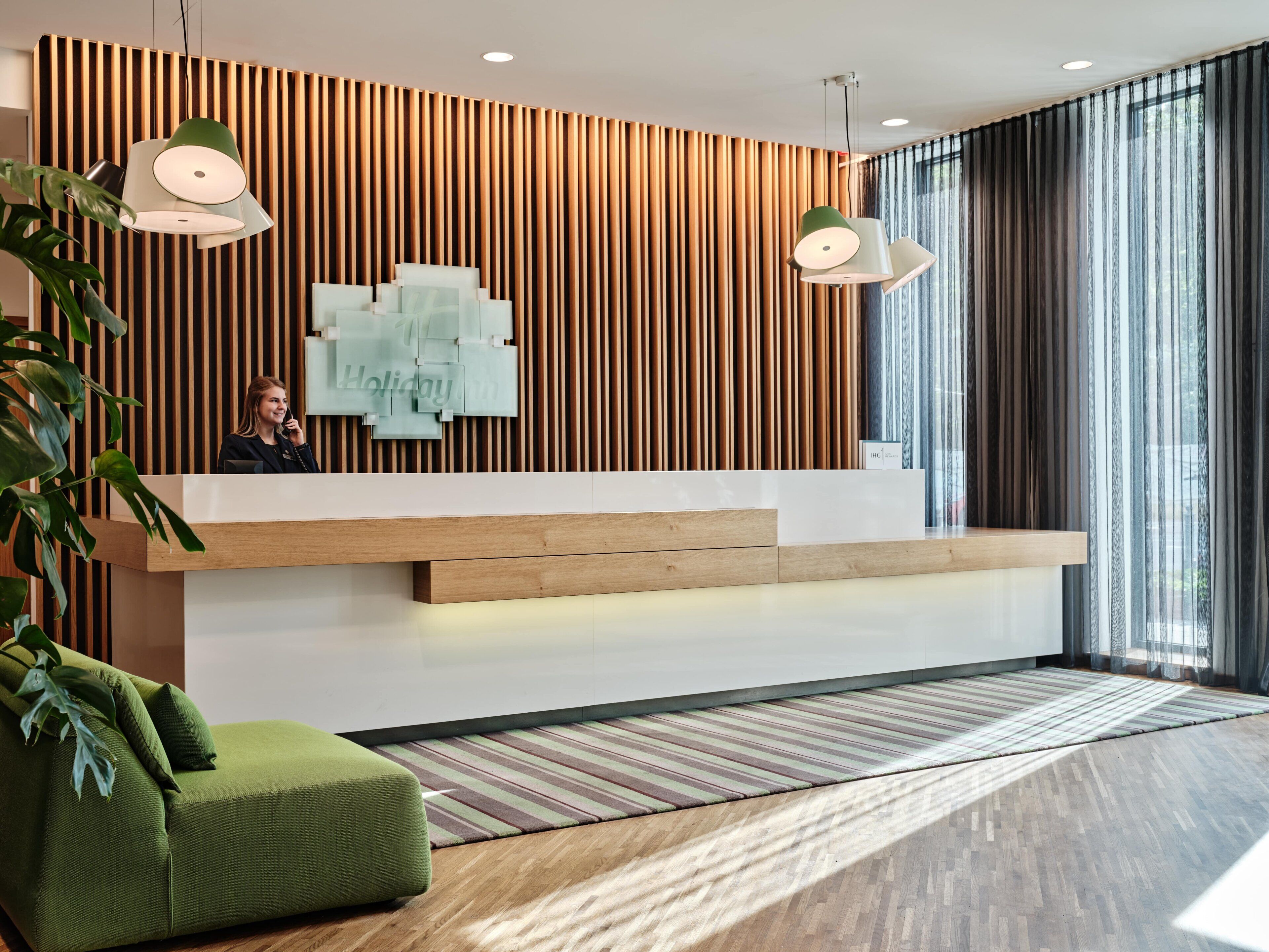 A modern reception desk with vertical wooden slats, a green upholstered seating area, and a large window with sheer curtains.