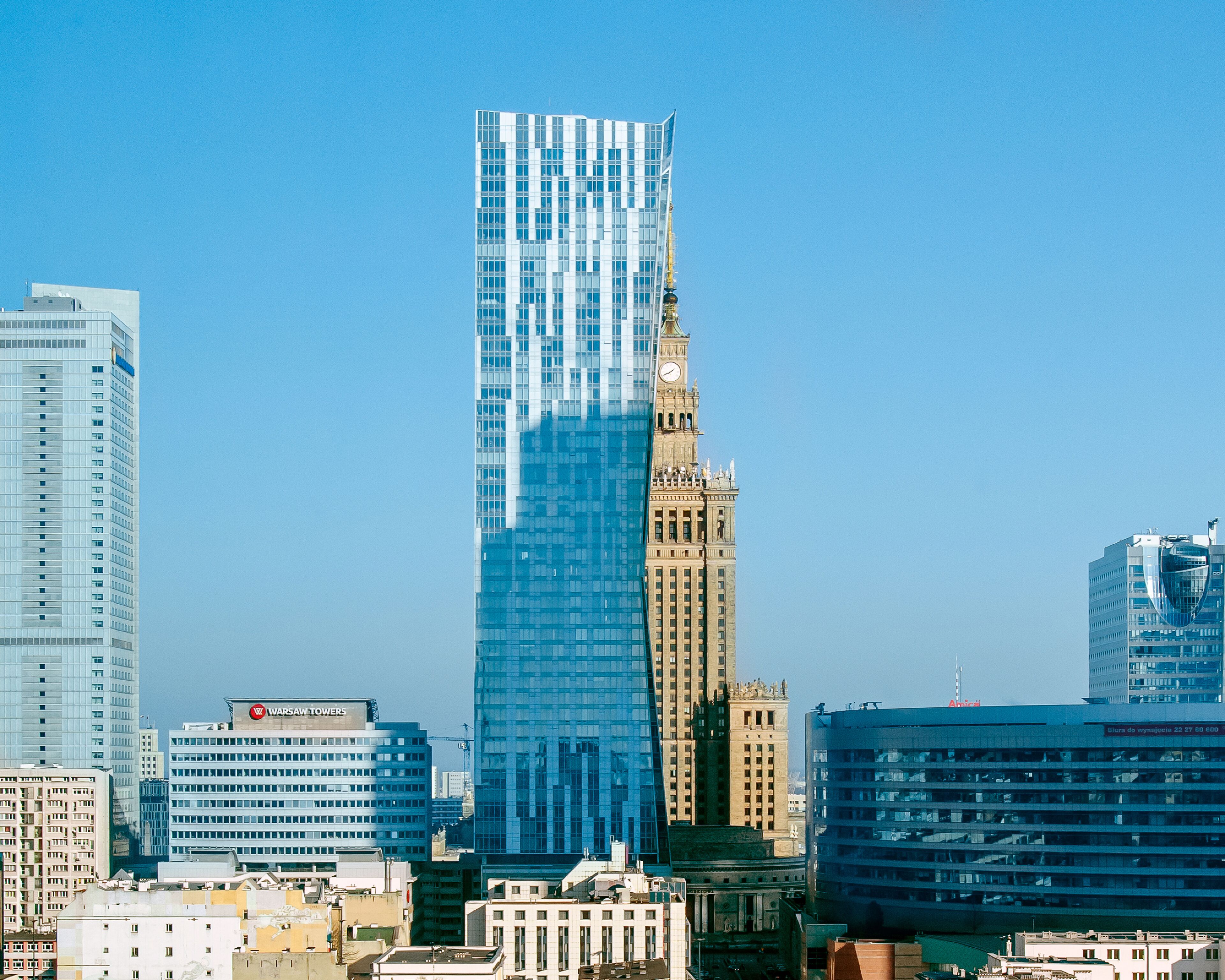 Un paysage urbain comprenant un imposant bâtiment moderne en verre, un bâtiment historique doté d’une tour horloge, ainsi que plusieurs autres immeubles de taille moyenne.