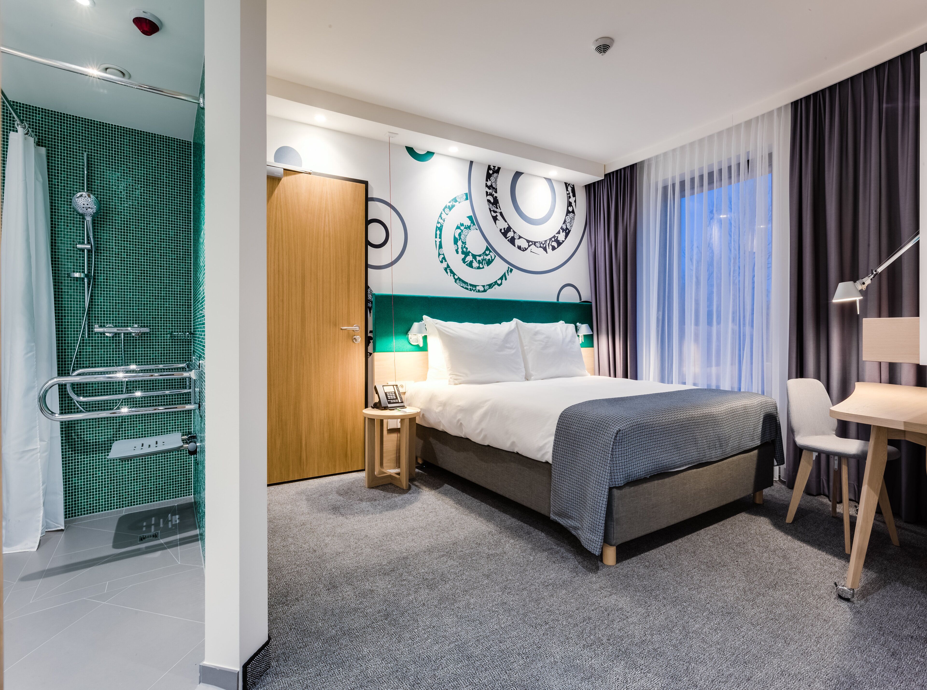 A hotel room with a bed, bedside table, chair, and a shower area with a towel rack.
