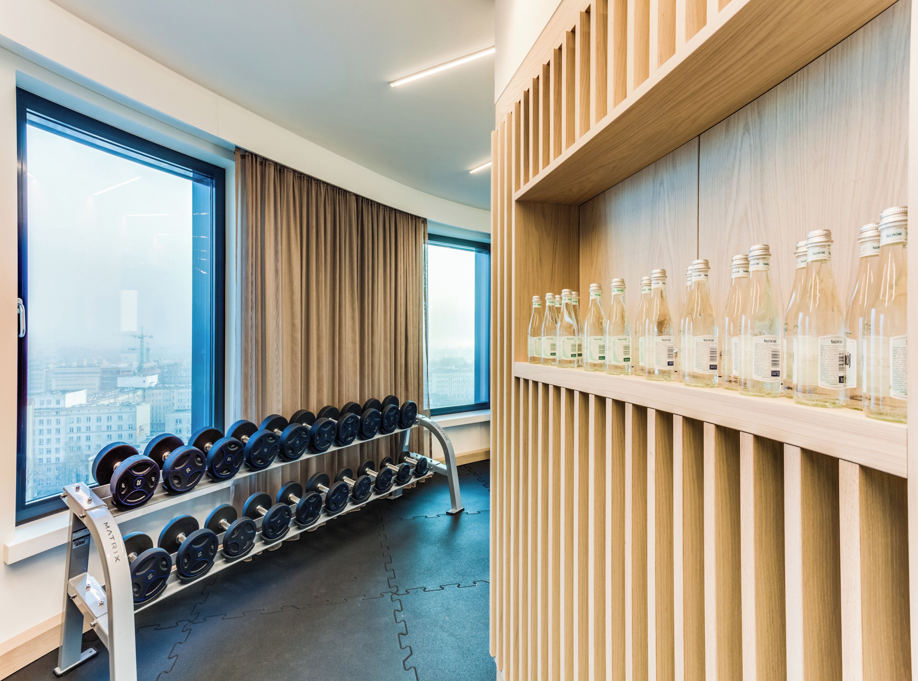 A gym with a rack of dumbbells, a shelf with water bottles, and a large window with a city view.
