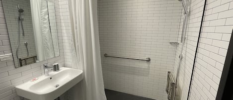 Une salle de bain avec des murs recouverts de carreaux blancs, un lavabo, une douche avec une porte en verre et une barre d’appui.