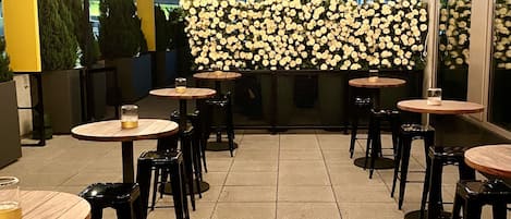 Un área de comedor al aire libre con varias mesas redondas y sillas negras, un muro decorativo de flores blancas y vistas de la ciudad de fondo.