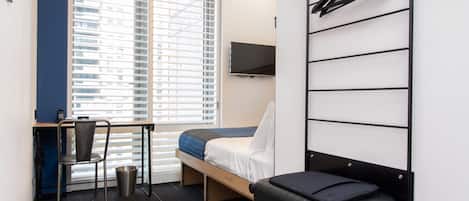Een kleine hotelkamer met een bed, een bureau, een stoel, een televisie en een bagageopbergruimte.