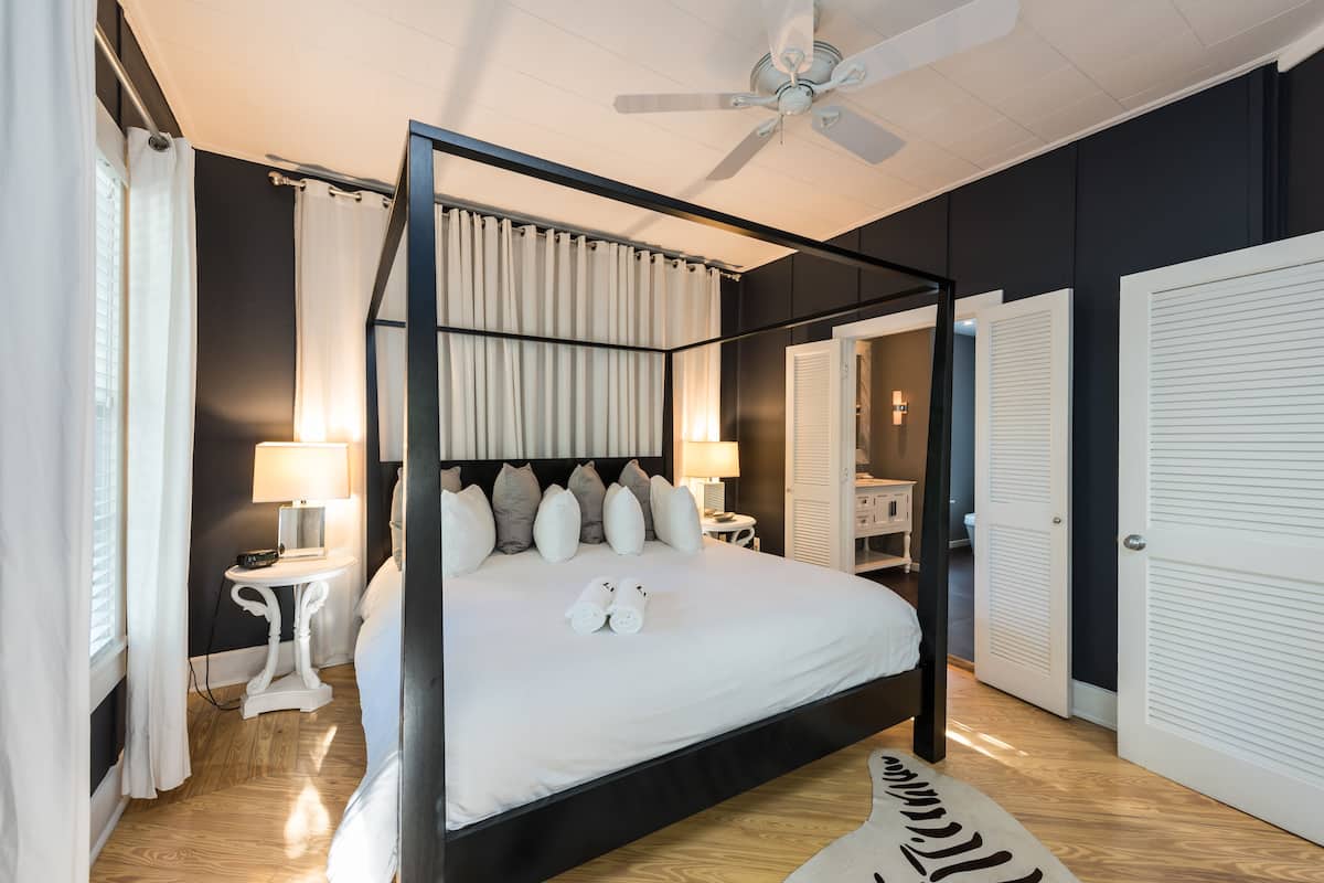 A four-poster bed with a canopy, bedside tables with lamps, a ceiling fan, and a view of a bathroom with a mirror and a wardrobe.