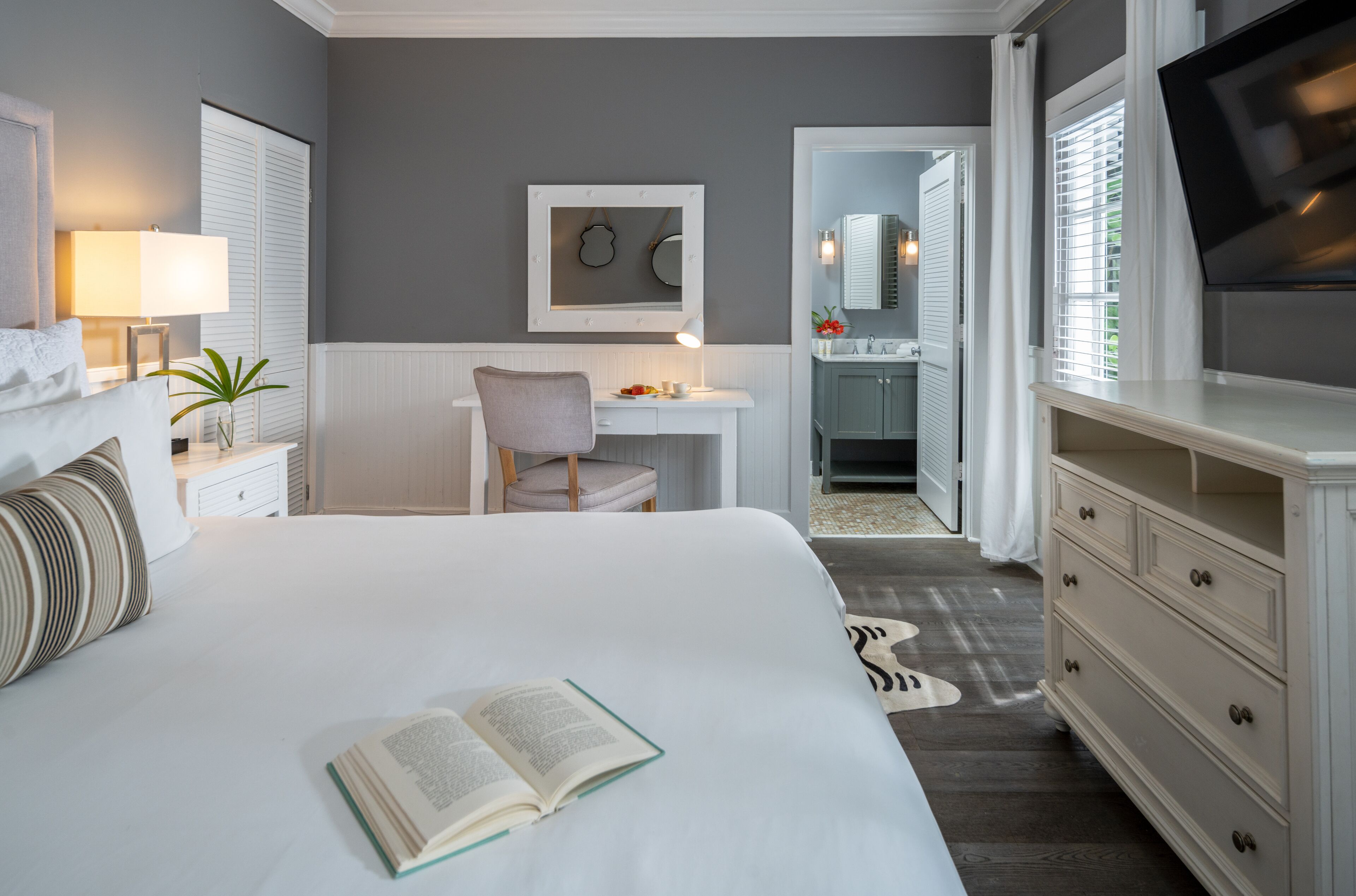 A bedroom with a large bed, a bedside table with a lamp, a chair, a desk with a chair, a mirror, a television, and a bathroom visible through an open door.