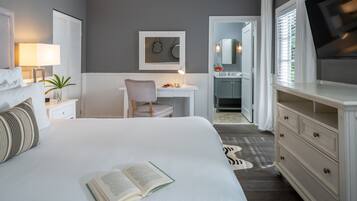 A bedroom with a large bed, a bedside table with a lamp, a chair, a desk with a chair, a mirror, a television, and a bathroom visible through an open door.