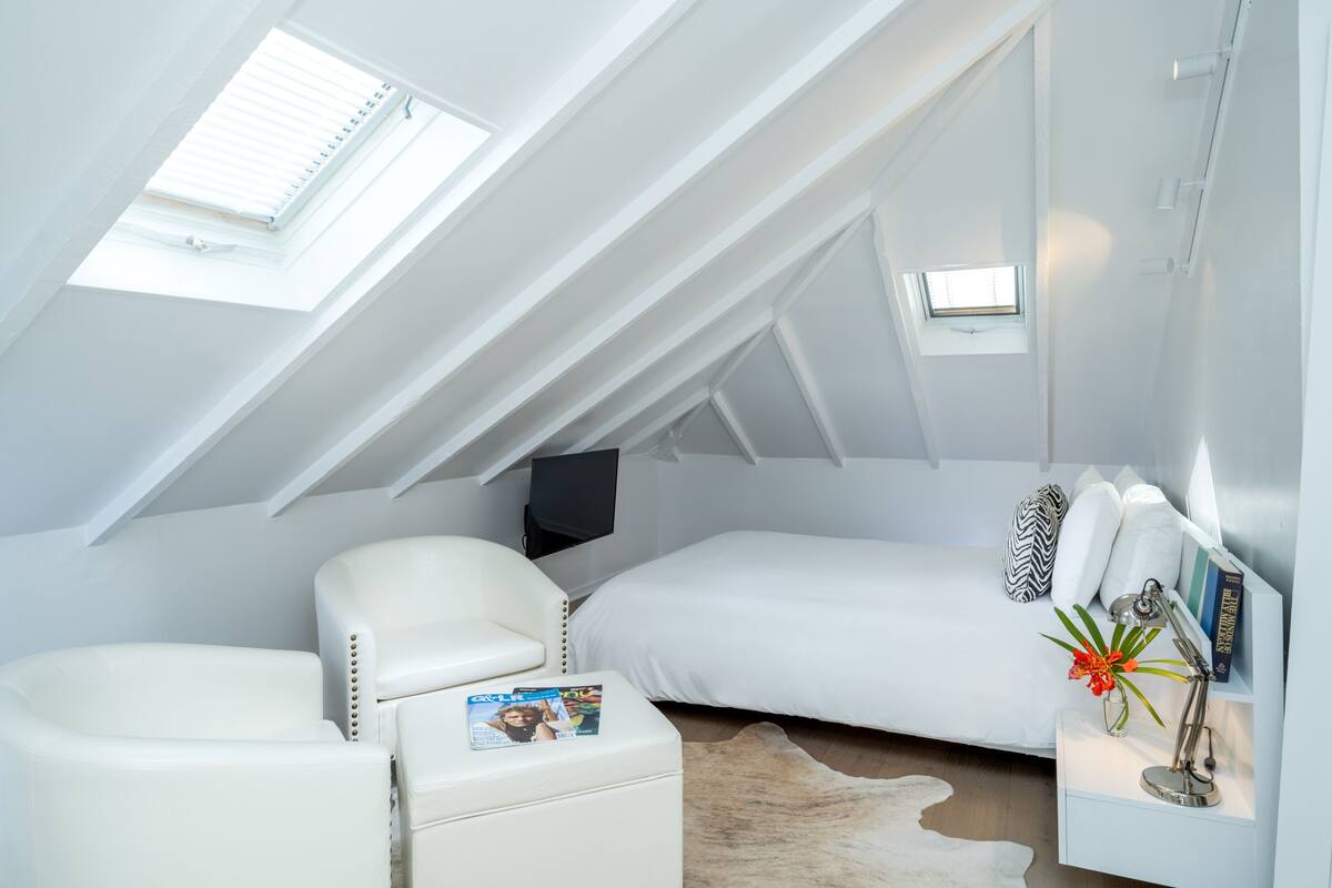 A modern loft bedroom with a sloped ceiling, a bed, a white leather armchair, a small bedside table with a lamp, and two skylights.