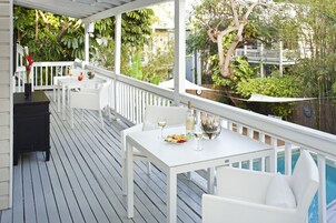 Un patio techado al aire libre con mobiliario blanco, una mesa de comedor dispuesta con comida y bebidas, y vistas a una piscina y follaje.