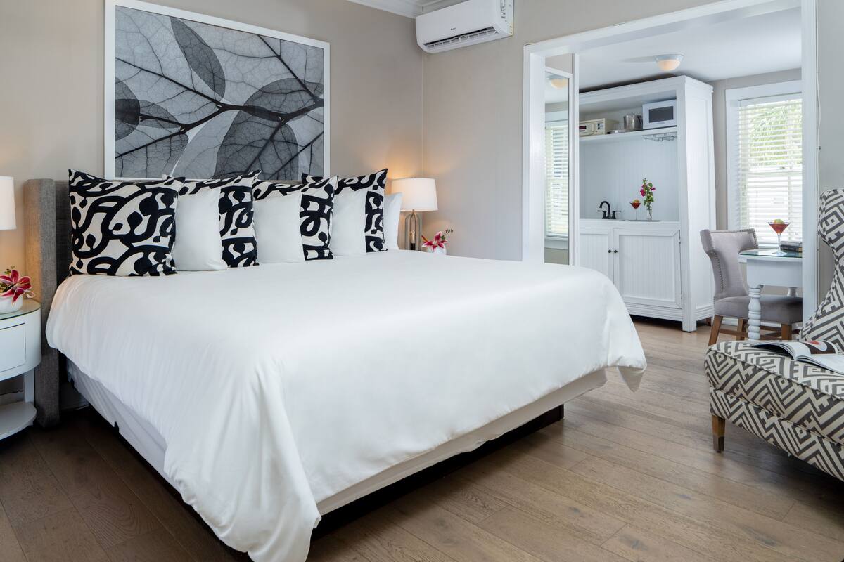 A neatly arranged bedroom with a large bed, bedside lamps, a patterned armchair, and a view into a connected room with a white cabinet and a chair.