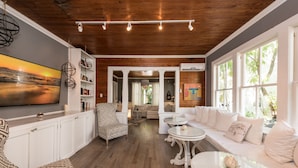 Amplia sala de estar con techo de madera, sofás blancos, un sillón estampado, una mesa de centro de cristal y un cuadro grande en la pared.