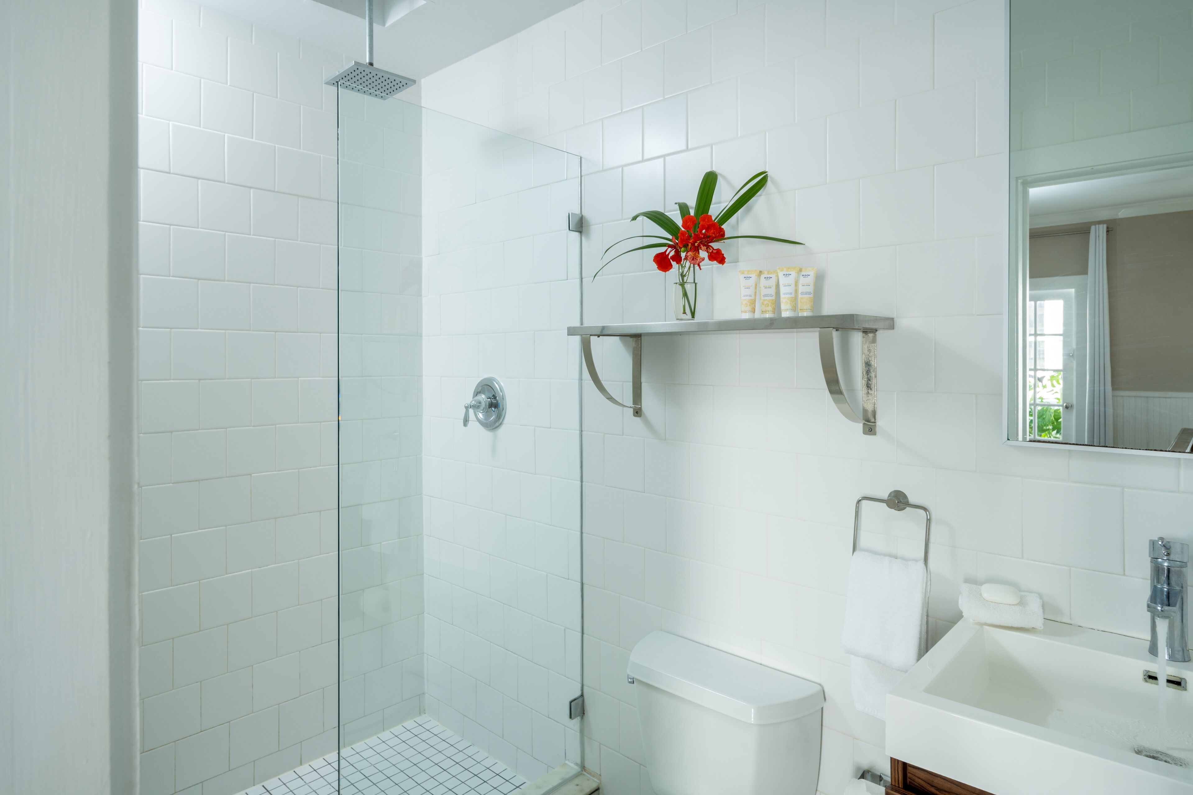 Un baño moderno con azulejos blancos, cabina de ducha de vidrio, inodoro, lavabo y estante con artículos de aseo.