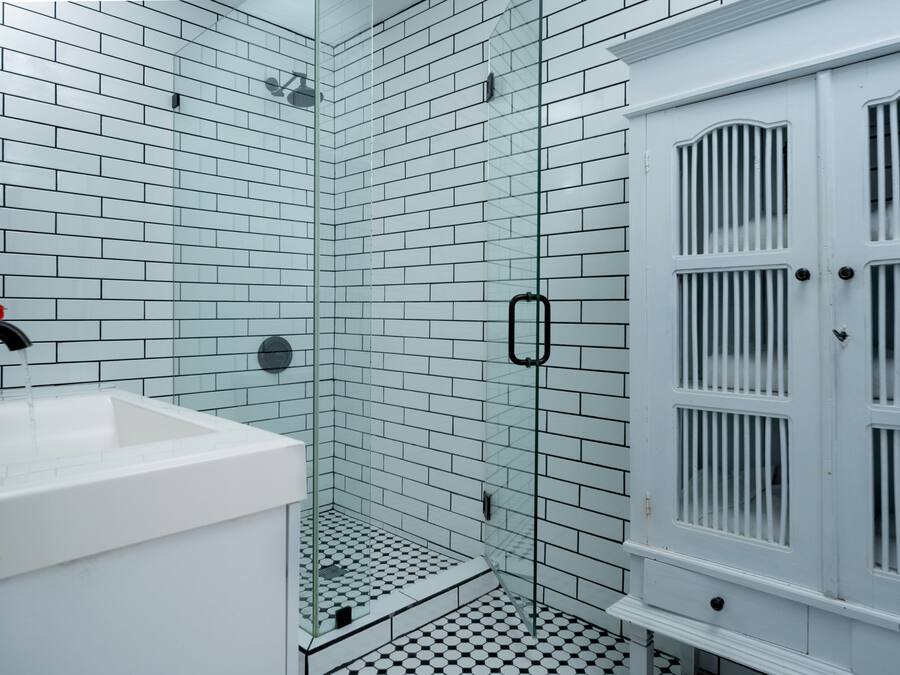 Un baño con azulejos blancos tipo metro, una cabina de ducha con mampara de vidrio y un mueble blanco con puerta.