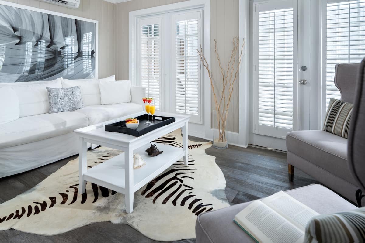 A modern living room with a white sofa, a white coffee table, and a grey armchair. There is a large abstract painting on the wall, a zebra-patterned rug, and a door with white shutters.