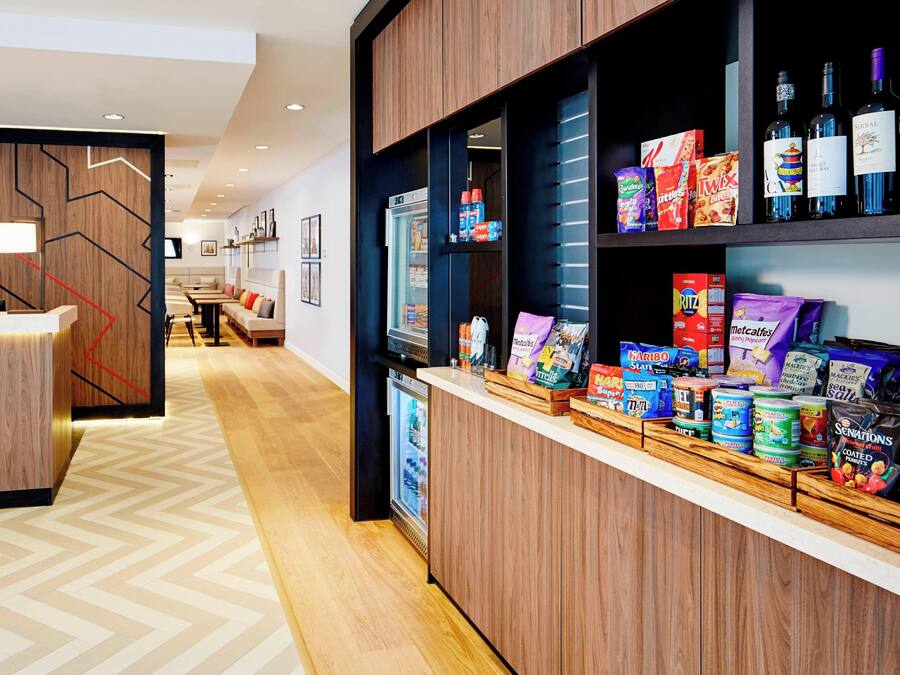 A hotel reception area with a wooden counter, a refrigerator, and shelves stocked with snacks, beverages, and toiletries.