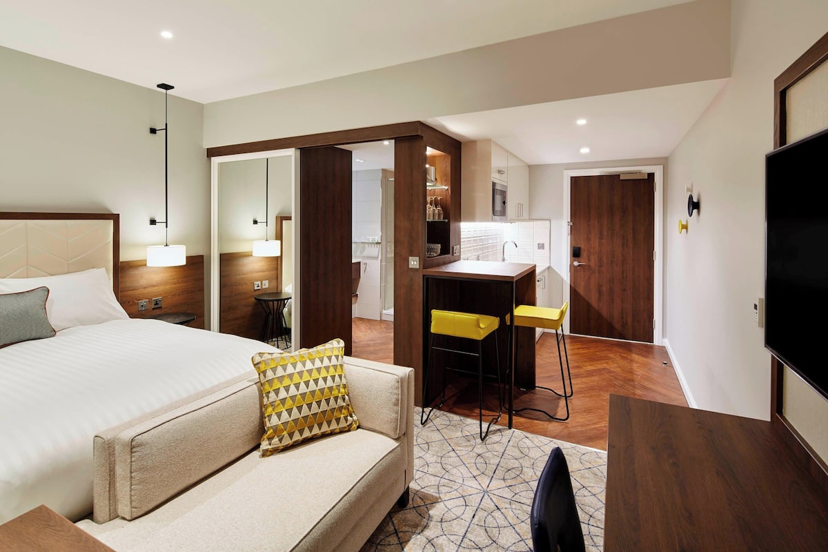 A modern hotel room with a bed, a sofa, a desk with two chairs, a TV, and a kitchen area.