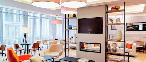 A modern hotel lobby with a seating area, a TV mounted on a wall, and a bookshelf with various items.