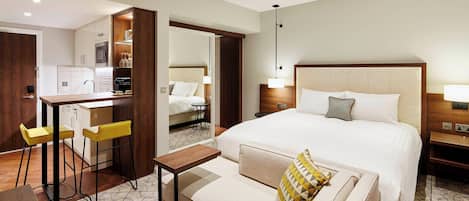 A hotel room with a bed, a sofa, a small table, a kitchen area, and a bathroom visible through an open door.