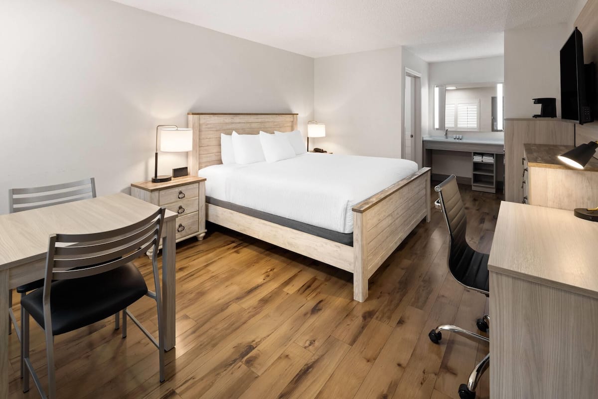 A hotel room with a wooden bed, a desk with a chair, a small table, a desk lamp, and a chair.