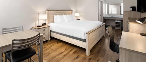 Habitación de hotel con una cama de madera, un escritorio con una silla, una mesita pequeña, una lámpara de escritorio y otra silla.