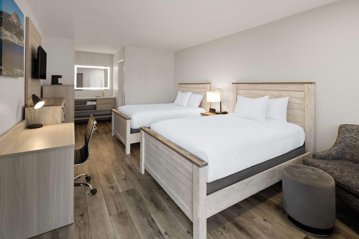 A hotel room with two beds, a desk, a chair, a small ottoman, and a bathroom visible in the background.