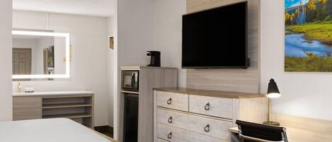 A hotel room with a bed, a desk with a chair, a dresser, a flat-screen TV, a microwave, and a painting on the wall.
