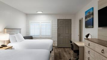 Quarto de hotel com duas camas, uma escrivaninha, uma cadeira, cômoda, televisão e janela com persianas.