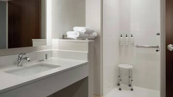 Un baño moderno con un lavamanos blanco, un espejo, una ducha con puerta de vidrio y un taburete pequeño.