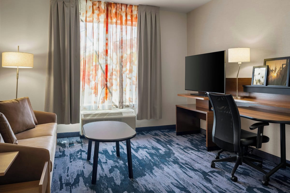 A hotel room with a sofa, a small table, a desk with a chair, a television, and a window with curtains.