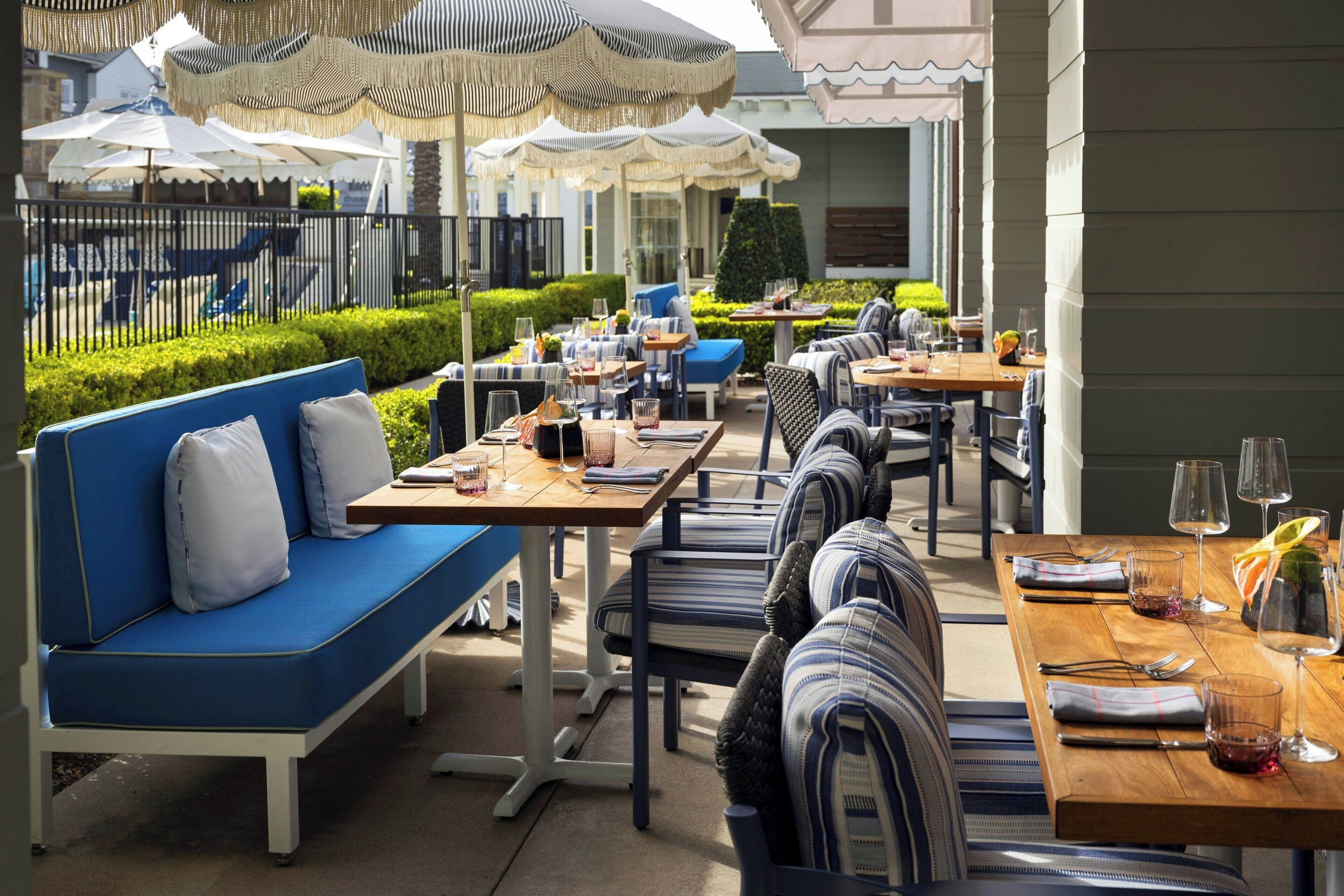An outdoor dining area with tables and chairs, umbrellas, and a pool in the background.