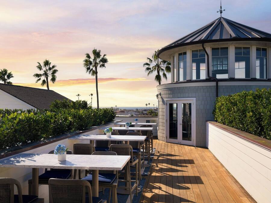 A rooftop patio with tables and chairs, palm trees, and a building with a distinctive roof.
