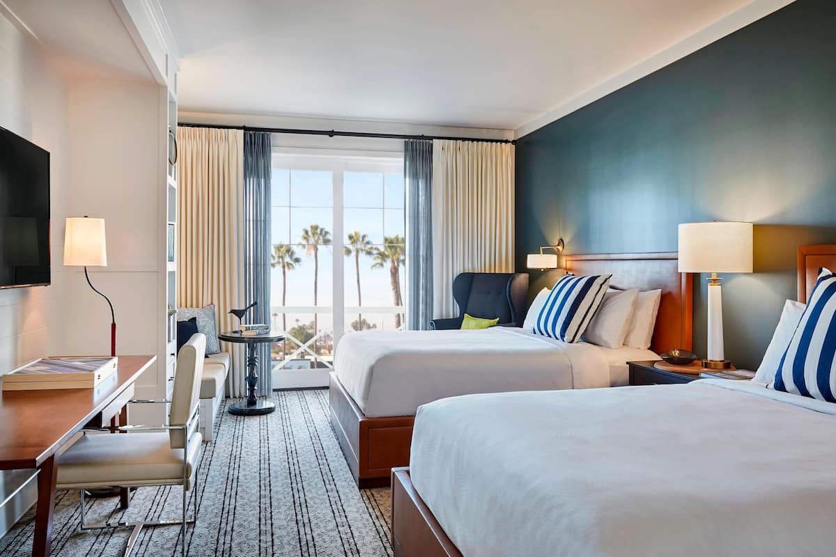 A hotel room with two beds, a desk, a chair, a TV, and a view of palm trees.