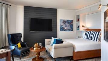 A hotel room with a large bed, a sofa, a chair, a TV mounted on the wall, and a small round table with glasses.