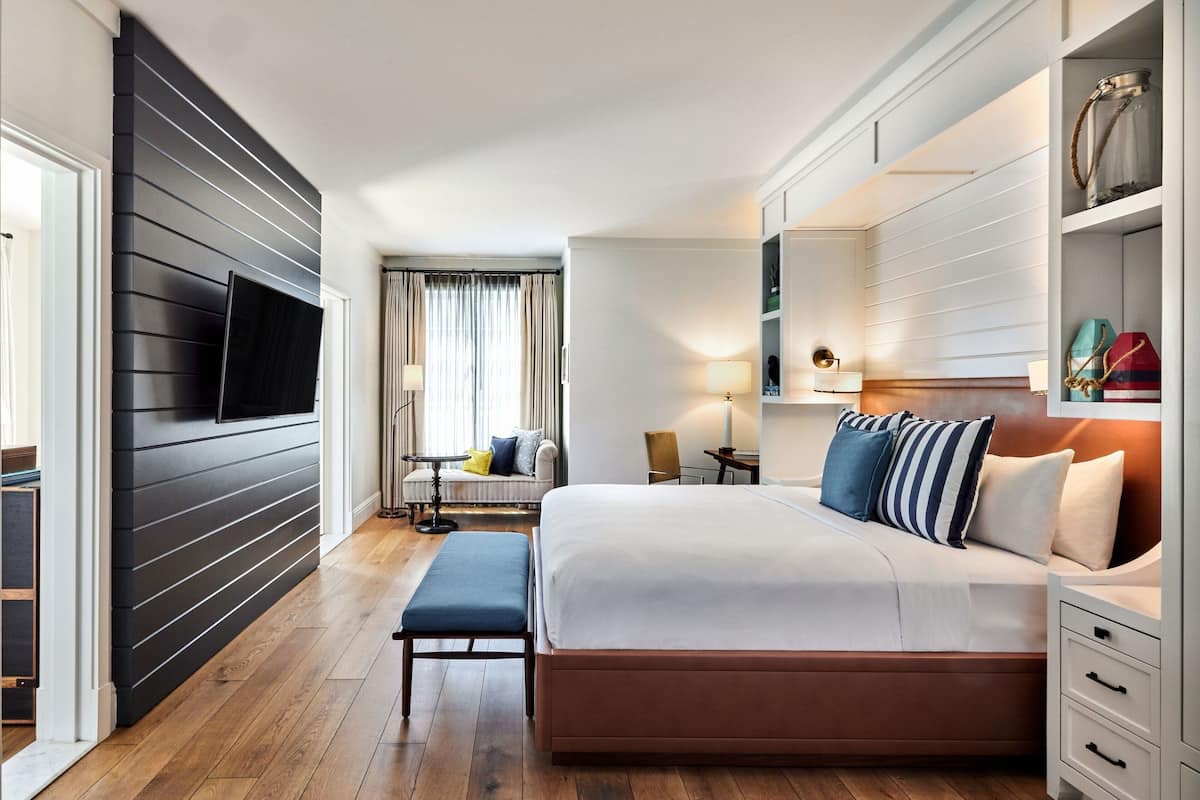 A modern hotel room with a large bed, a flat-screen TV mounted on the wall, a bedside table with a lamp, and a view of the hallway and another room.